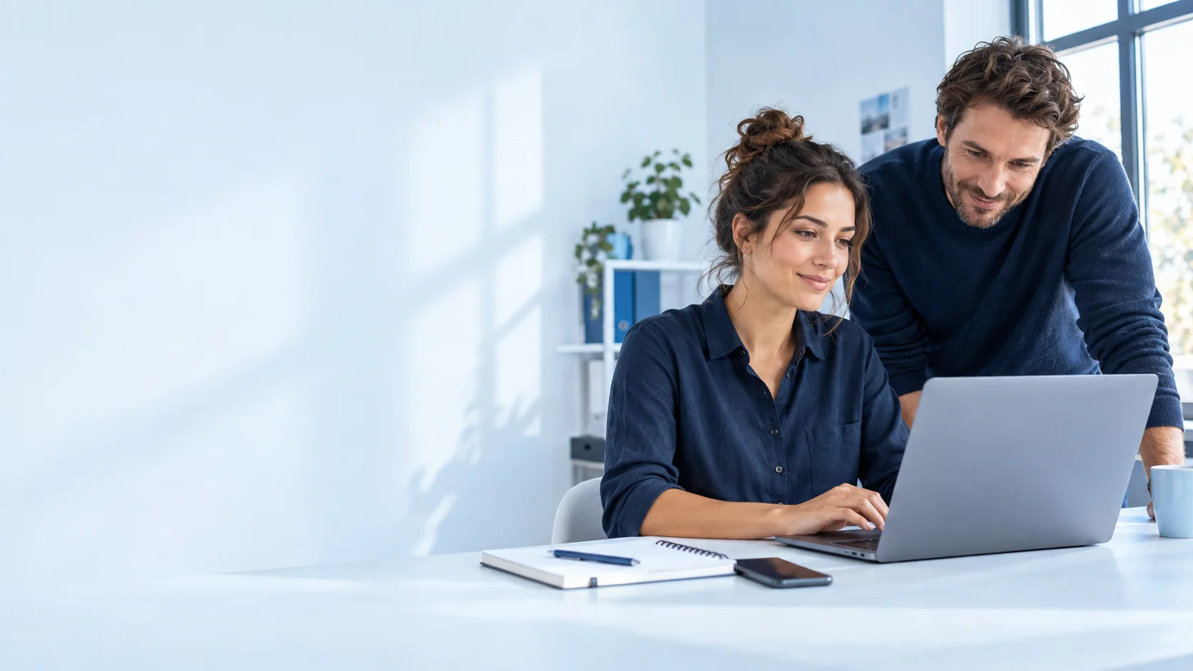 Two people working confidently together at a laptop in a bright, blue-white workspace. Illustrative artwork.